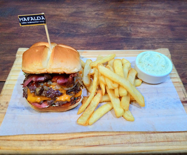 Hamburguesa de Mafalda con queso y bacon, acompañada de papas fritas y salsa blanca en tabla de madera.