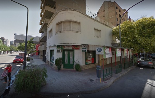 Edificio esquinero con fachada blanca y verde, puerta principal abierta, árboles y coches estacionados frente a Tomasino.