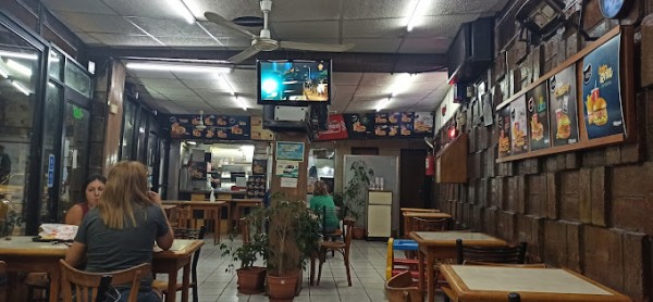 Interior de un bar con mesas, sillas, televisión encendida y paredes decoradas con carteles publicitarios.