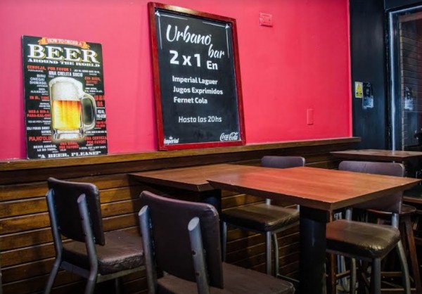 Interior de Urbano bar con mesas marrones, sillas altas y pared roja con carteles de cerveza y promociones.