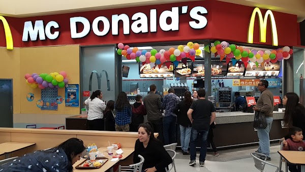 McDonald's con gente comiendo y haciendo fila. Decoración con globos coloridos. Ambiente familiar y animado.