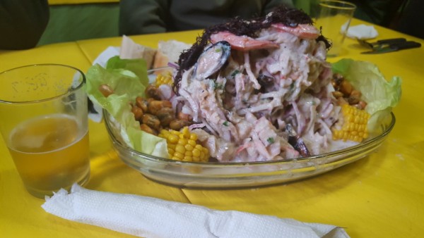 Plato de ensalada con mariscos y maíz en mesa amarilla. Bebida y servilleta al lado. Restaurante 