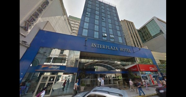 Entrada del Interplaza Hotel con fachada azul, gente caminando y tiendas alrededor. Edificio moderno en ciudad.