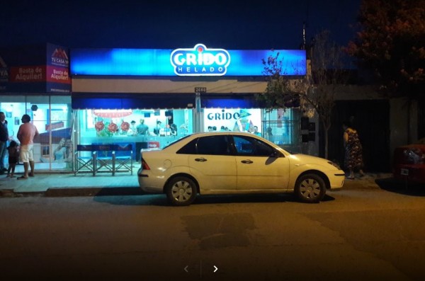 Noche en Grido Helado, Bº Yofre. Local iluminado con gente adentro y auto blanco estacionado frente. Árbol y cartel rojo al costado.