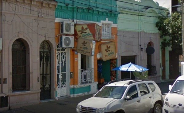 Edificios coloridos con balcones y puertas arqueadas en Ron Damón. Autos estacionados frente a locales comerciales.