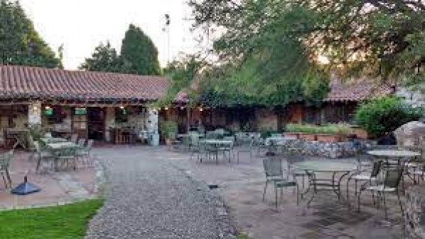 Rancho Viejo: patio con mesas y sillas bajo árboles frondosos, edificio de techo rojo al fondo. Ambiente tranquilo y acogedor.